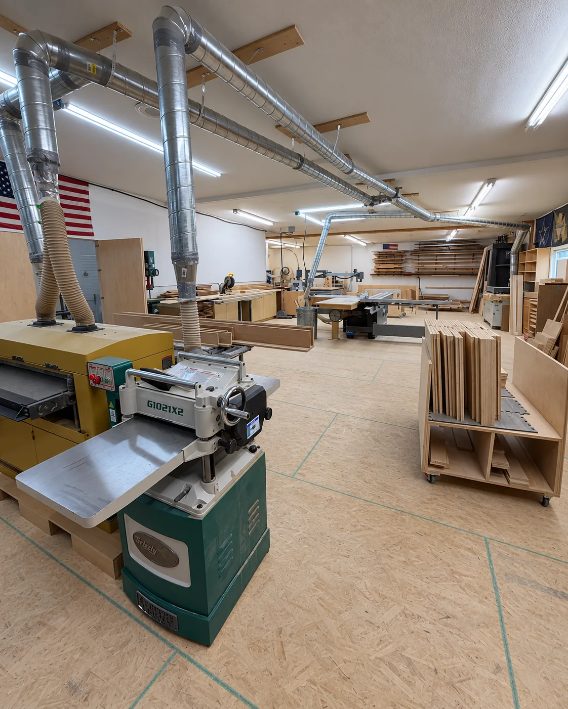 The Ringgold, Georgia workshop: a planer and a sander in the foreground under overhead dust-collection ducting, a rolling cart of cut panels to the right, and a table saw and material racks along the far wall.